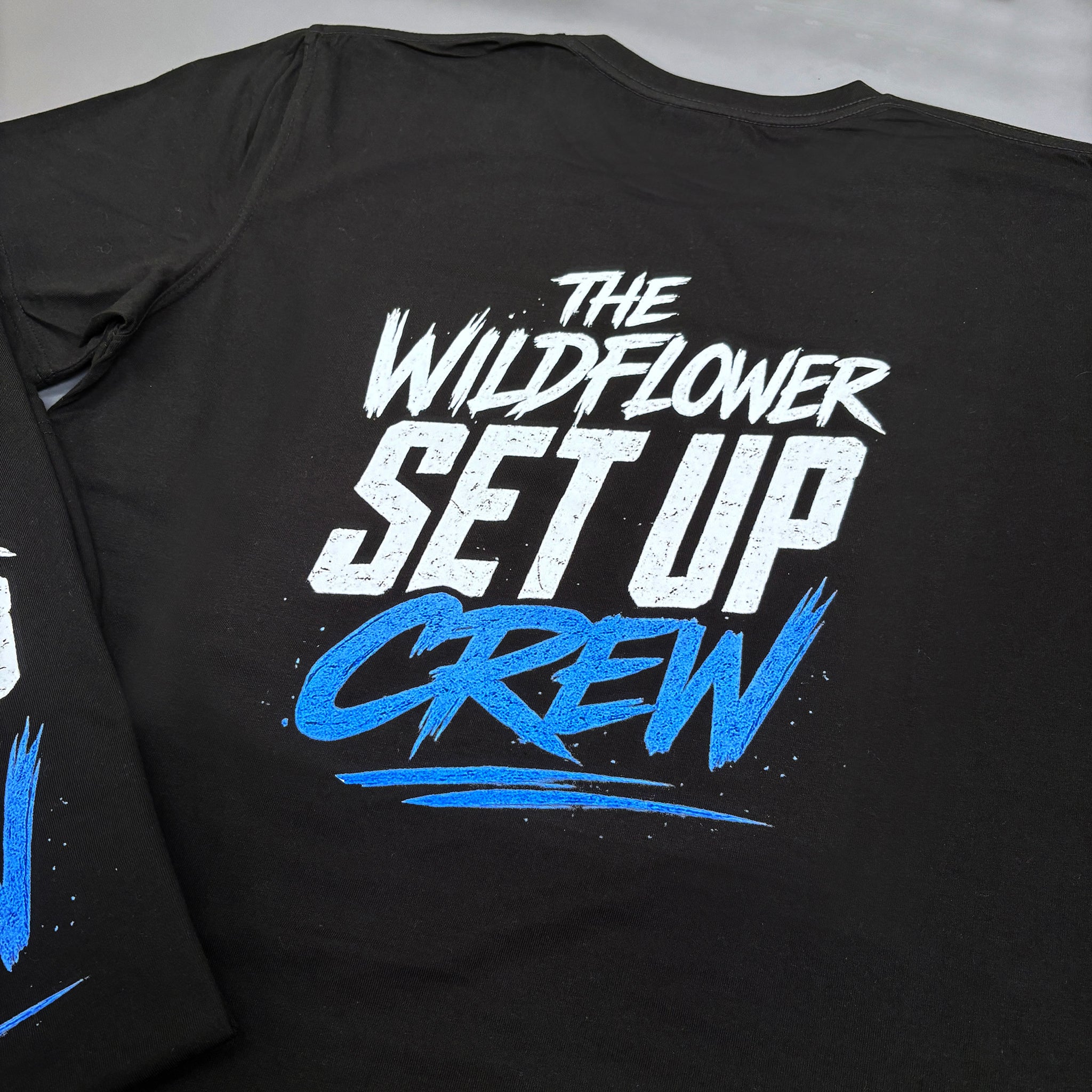 Black printed t-shirt with 'The Wildflower Set Up Crew' text in white and blue on a black background